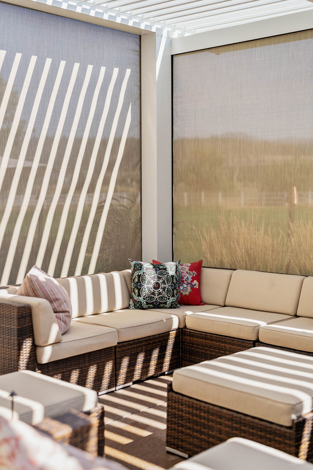 Sectional couch with dappled light streaming through an opened white aluminum StruXure louvered pergola above with tan screens from MagnaTrack lowered and visible behind the couch creating a screened in porch feel
