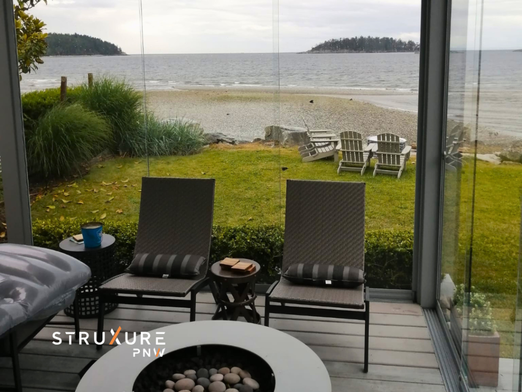 Inside looking out view from a patio enclosed with glass partitions showing a fire pit and two chairs inside with a view of a lake shoreline and stormy Pacific Northwest sky through the glass